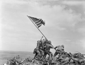 Historic black-and-white photograph of six U.S. Marines raising an American flag on a tall flagpole atop a rocky hill during World War II’s Battle of Iwo Jima. The soldiers work together, leaning forward and pushing the pole into the ground, with debris and rough terrain beneath them and a cloudy sky in the background.