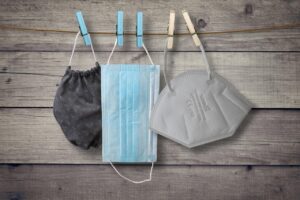Three types of face masks hanging on a clothesline with clothespins against a wooden background. From left to right: a reusable black patterned cloth mask, a blue disposable surgical mask, and a white N95 respirator mask.